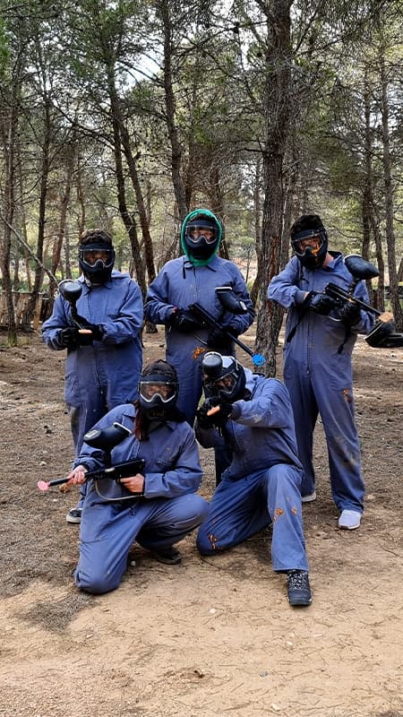 Jugador de paintball protegido con máscara y mono apuntando con su marcadora en un escenario exterior con obstáculos.